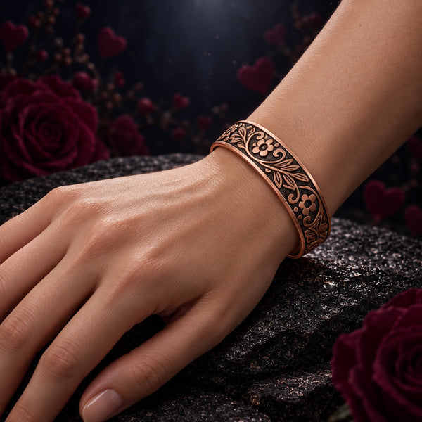 Venus Code copper bracelet with flower and leaf motifs, worn on a hand model’s wrist with burgundy roses and warm heart shaped lights in the background.